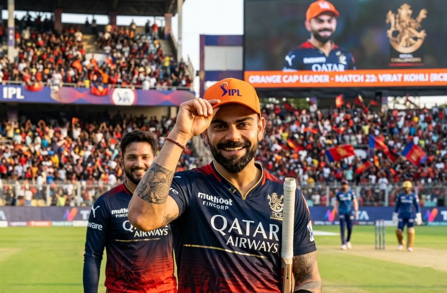 A high-resolution photograph of Virat Kohli on a cricket field, smiling and tipping an orange IPL cap toward the camera. He is wearing the Royal Challengers Bengaluru (RCB) navy and red jersey with sponsor logos "Qatar Airways" and "Muthoot FinCorp." In the background, a blurred stadium crowd and a large digital screen are visible, with the screen displaying his portrait and the text "Orange Cap Leader - Match 23: Virat Kohli (RCB)." A teammate stands smiling behind him to the left.